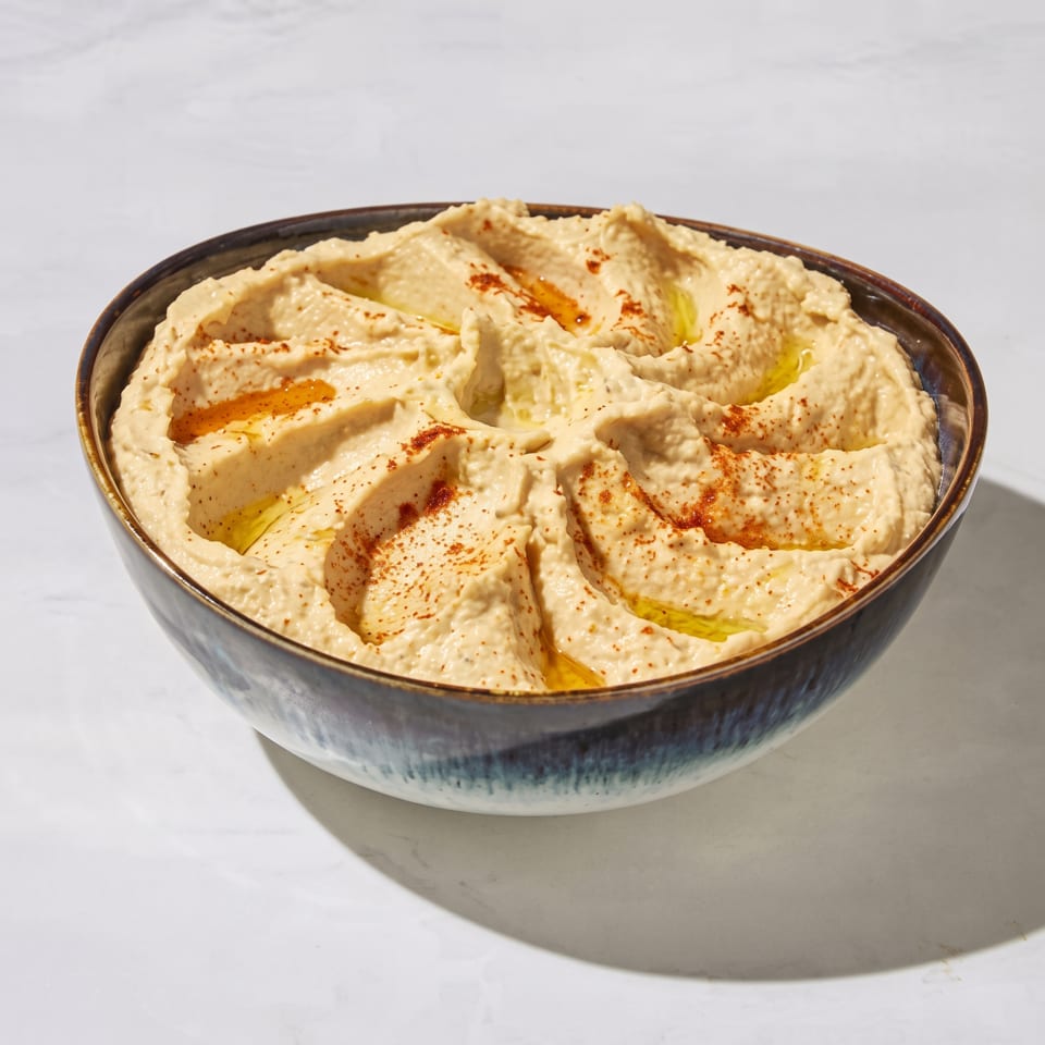 Bowl of creamy hummus drizzled with olive oil and sprinkled with paprika on a white countertop.