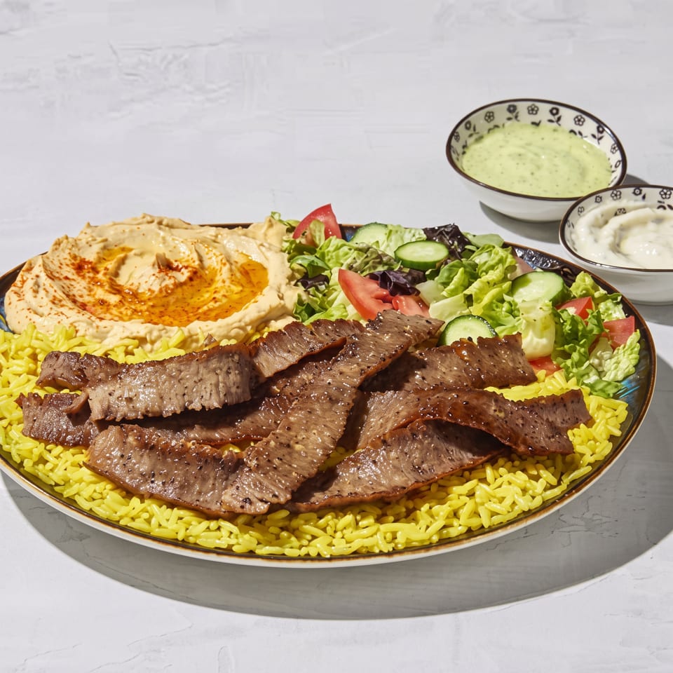 Platter of grilled meat over yellow rice with hummus, salad, and bowls of creamy sauces.