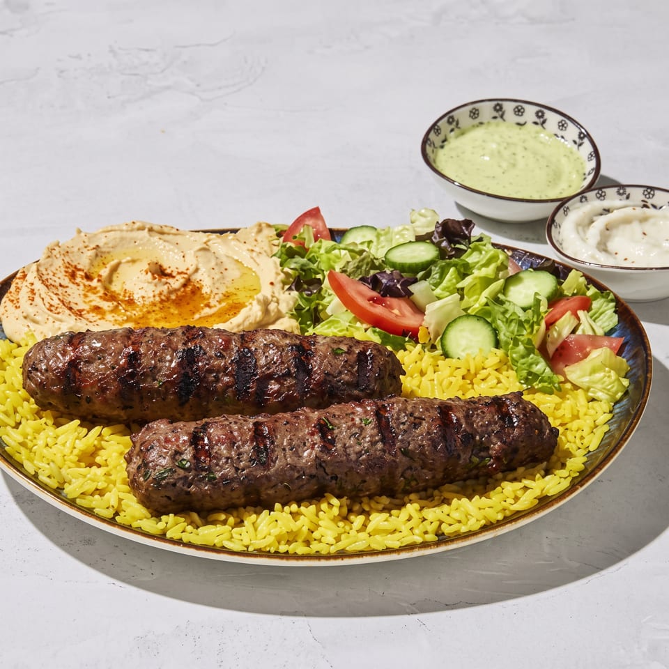 Two grilled kofta kebabs served over yellow rice with hummus, salad, and bowls of creamy sauces.