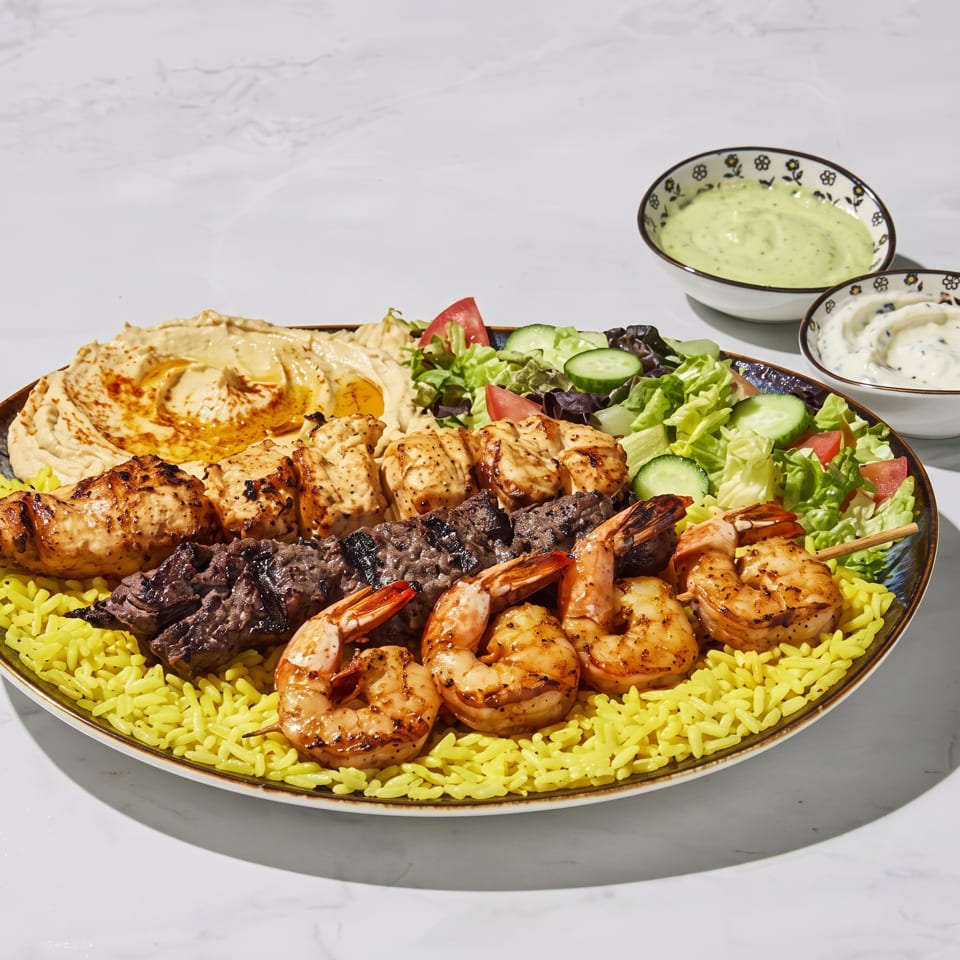 Mixed grill platter with grilled chicken, beef, shrimp, yellow rice, hummus, salad, and creamy dipping sauces.